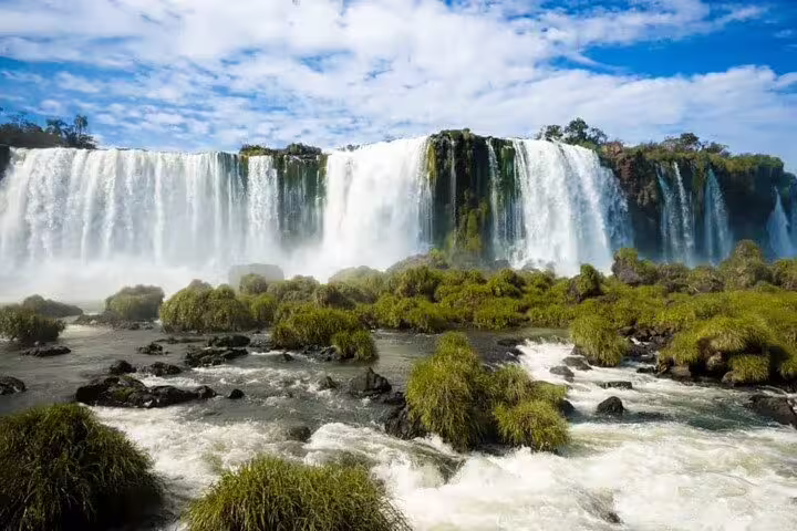 Stunning view of Iguazu Falls cascading over lush greenery under a bright blue sky, perfect for a thrilling boat tour.