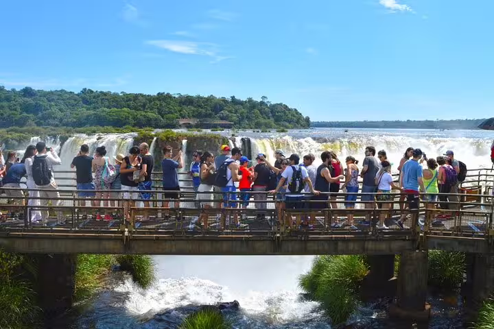 Tourists on a scenic walkway admire the panoramic views of Iguazu Falls under a clear blue sky during the guided tour.
