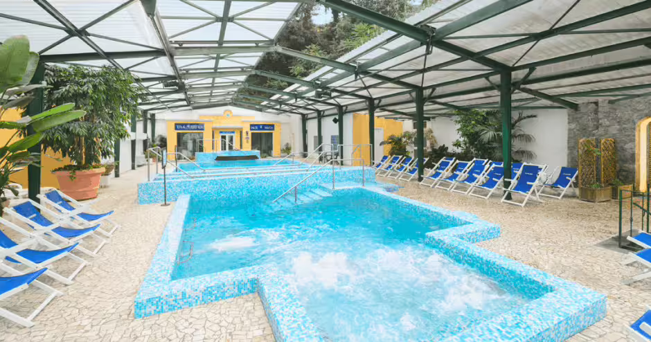 Piscine termali coperte del Parco Castiglione a Ischia, con idromassaggio, lettini blu e vegetazione mediterranea.
