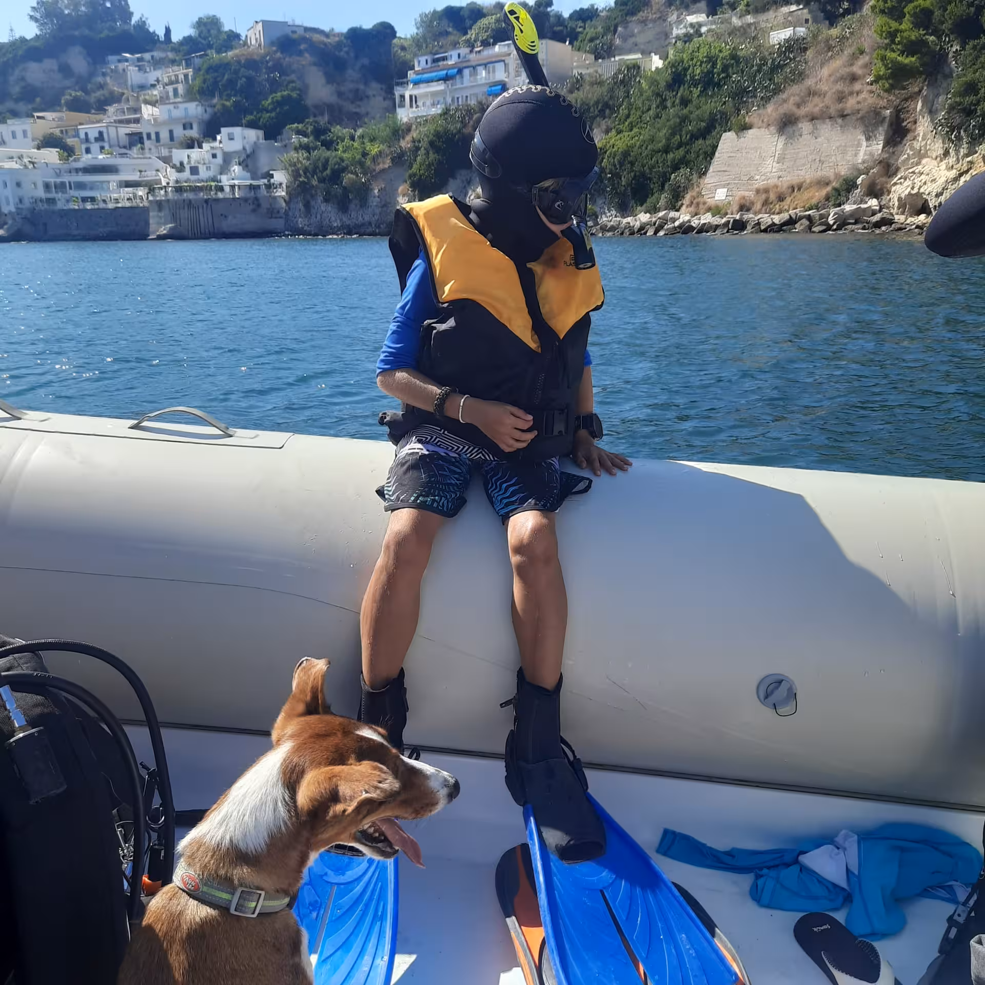 A young kid all dressed up in snorkeling equipment, with a life jacket, getting ready to go in the water from the boat, next to his dog..