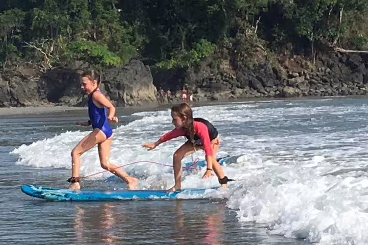 Two kids enjoying surf lessons on gentle waves at Manuel Antonio beach, surrounded by lush greenery.