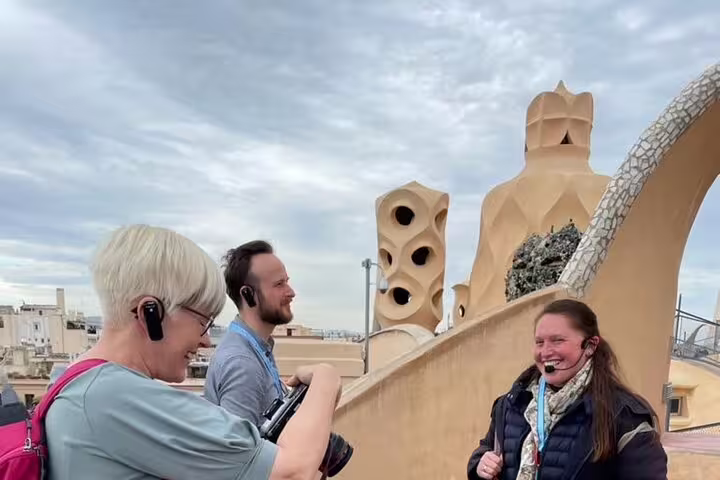 Small group with headsets enjoying a guided morning tour on La Pedrera rooftop in Barcelona, with Gaudí chimneys behind them