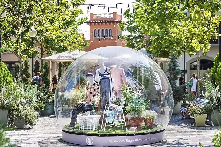 Stylish mannequin display in a glass dome at La Roca Village, surrounded by lush greenery and shoppers.