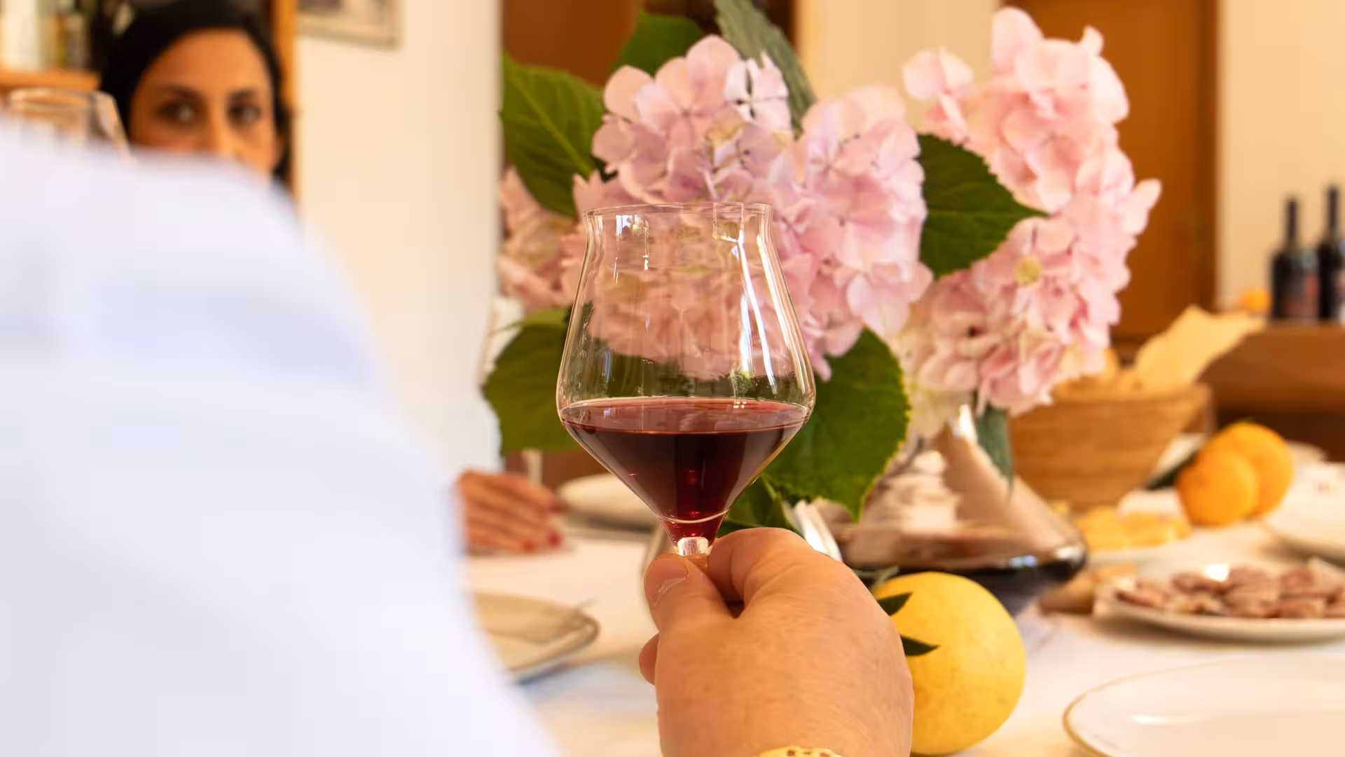 A person holding a glass of red wine at a Laconi winery tasting, surrounded by vibrant pink hydrangeas and fresh fruits.