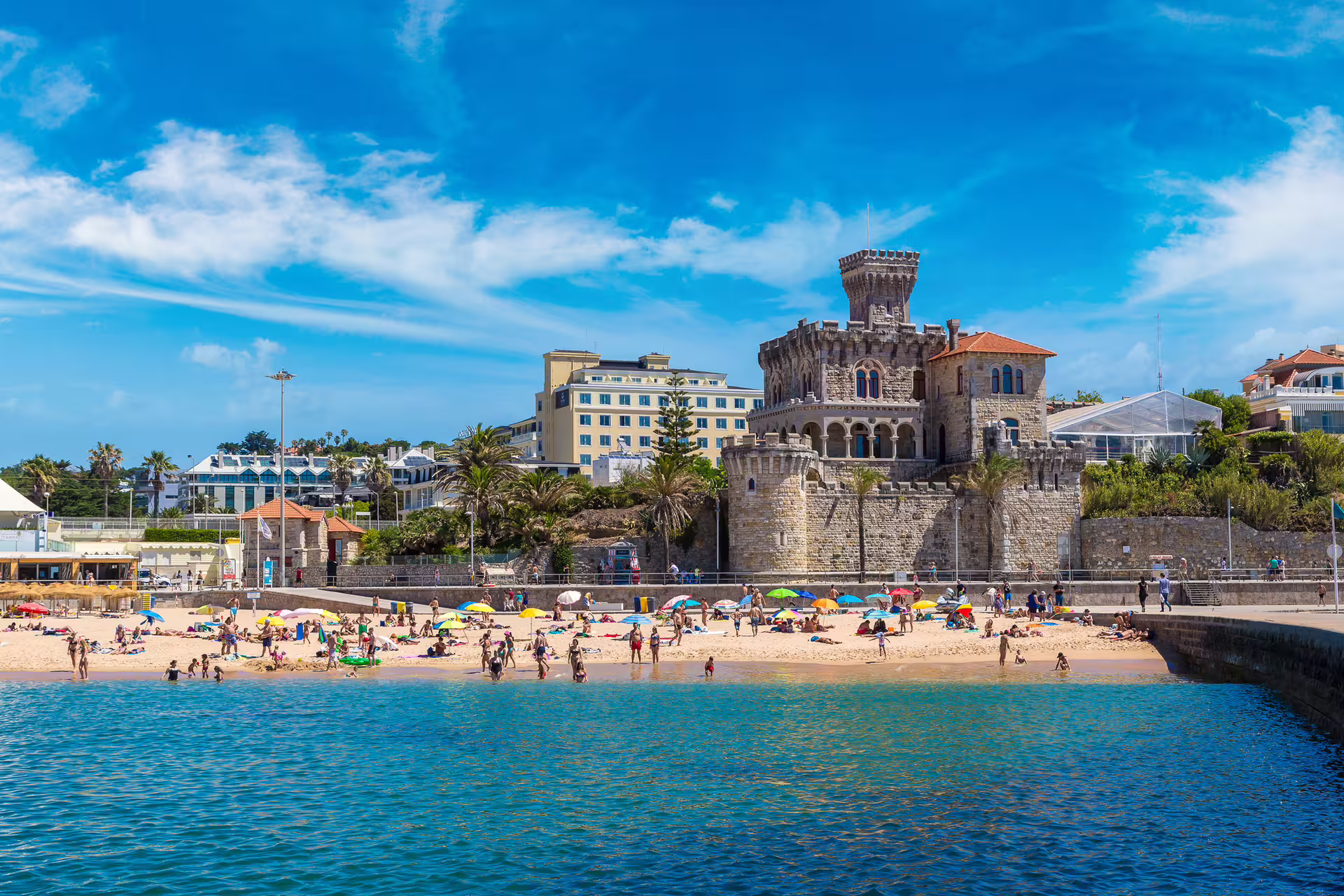 Beachgoers enjoy a sunny day at a scenic beach near a historic castle in Estoril, Portugal, part of the Lisbon shore excursion.