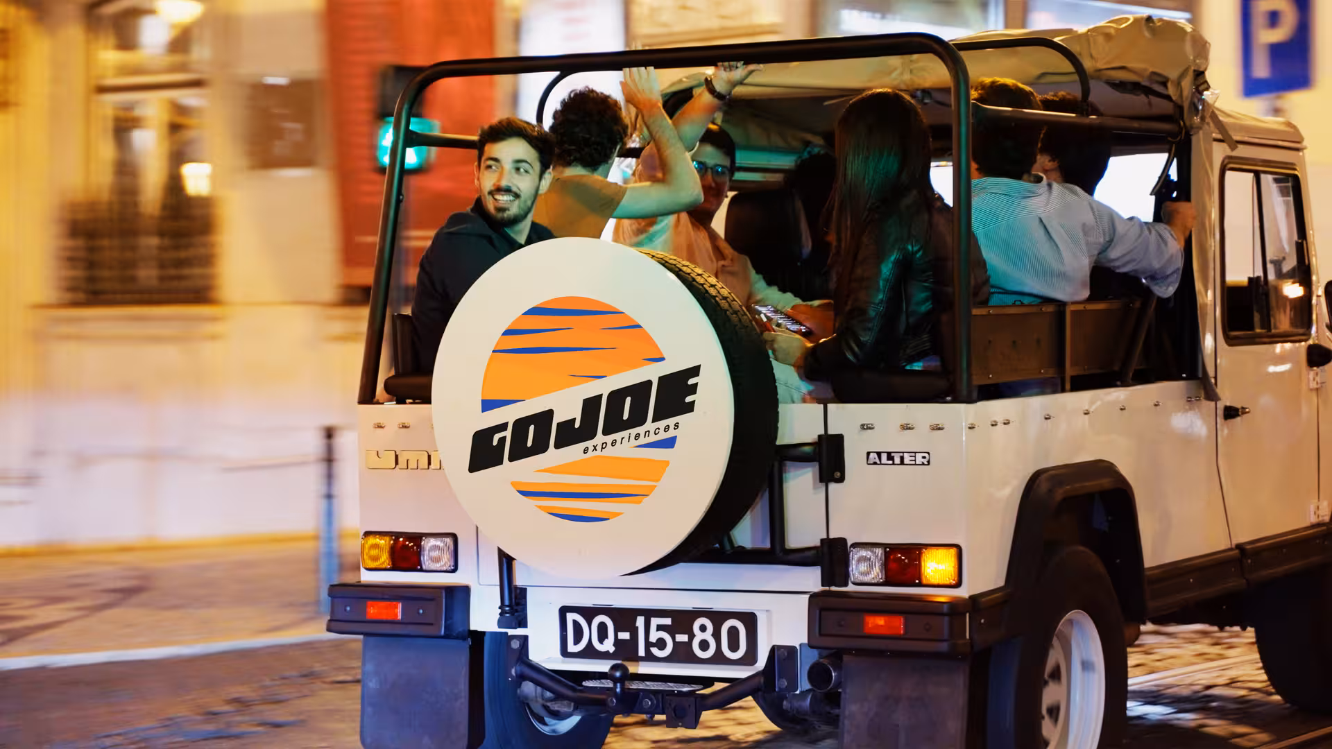 Friends riding a vintage Jeep at night in Lisbon, waving near a landmark on sunset-to-night tour