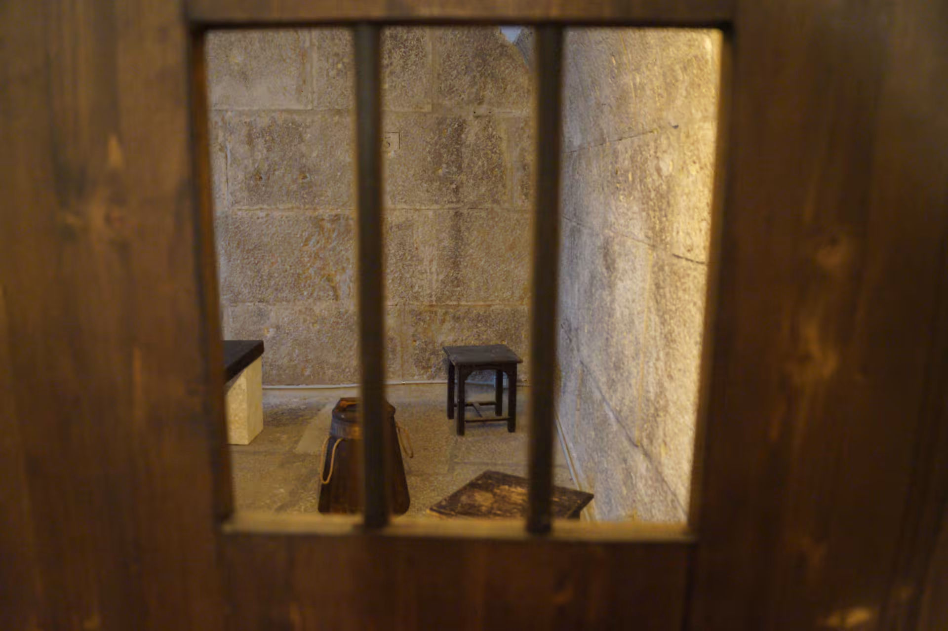View through barred door into a sparse cell at Lords of The Night, featuring stone walls and minimal furnishings.