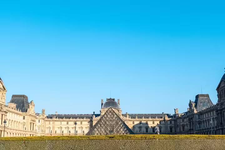 View of the iconic Louvre Pyramid with the historic museum facade under a clear blue sky in Paris, France.