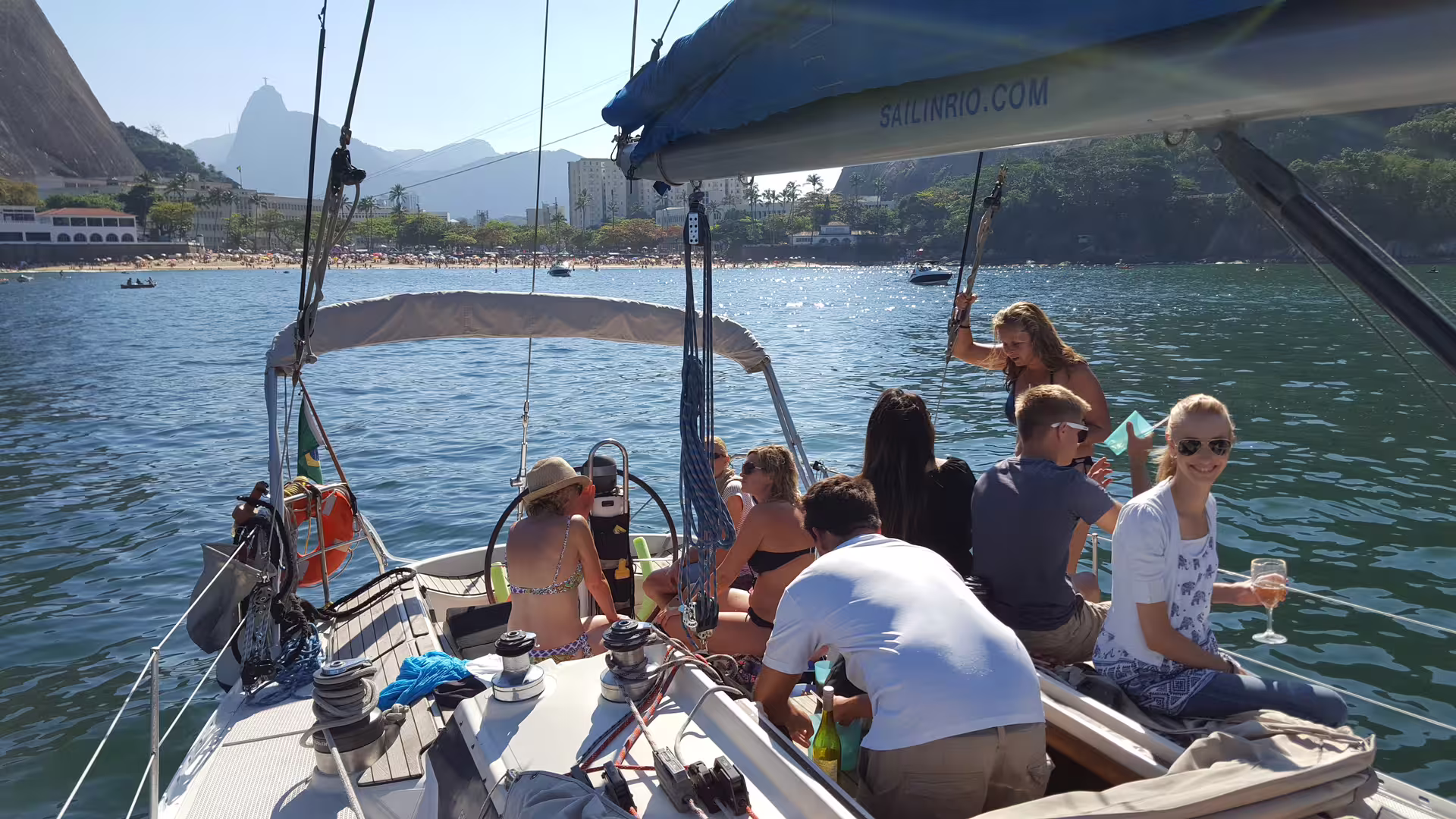 Group of friends enjoying a vibrant day on Makani Maikai sailboat with scenic city and mountain backdrop.