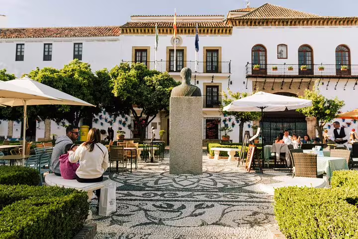 Scenic plaza in Marbella Old Town featuring a statue, outdoor cafes, and lush greenery under a clear blue sky.