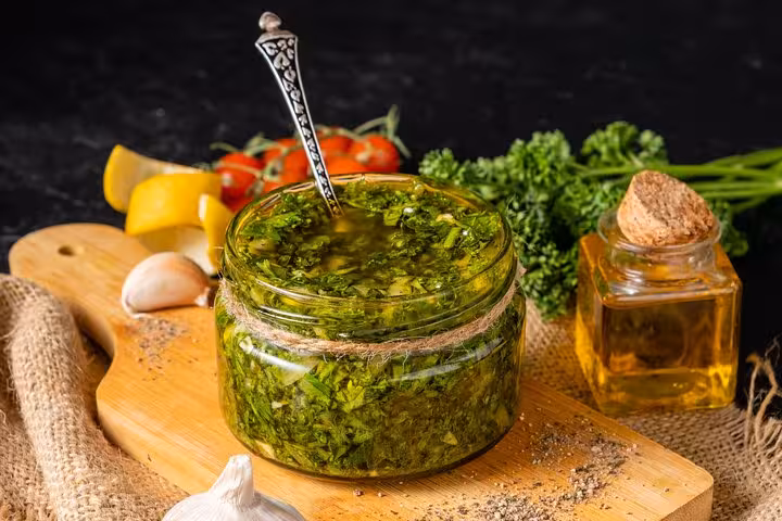 Fresh homemade chimichurri sauce in a glass jar with a spoon, accompanied by garlic, olive oil, and herbs on a wooden board.