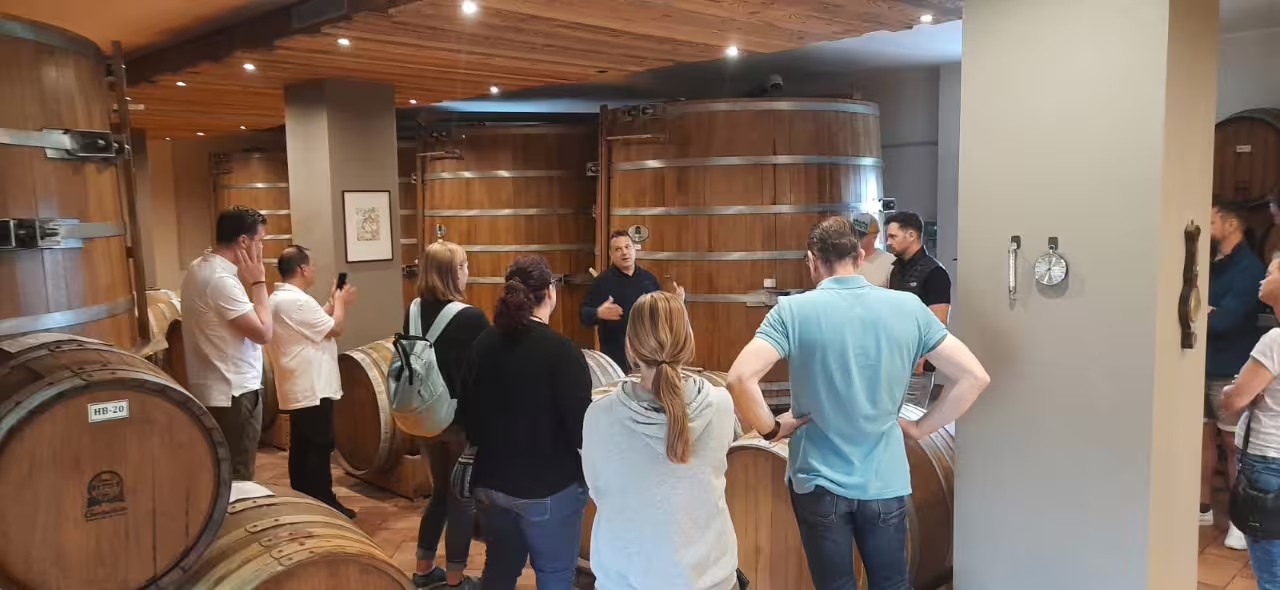Tour group gathers in Modena's balsamic vinegar cellar, listening intently to a guide's explanation amid wooden barrels.