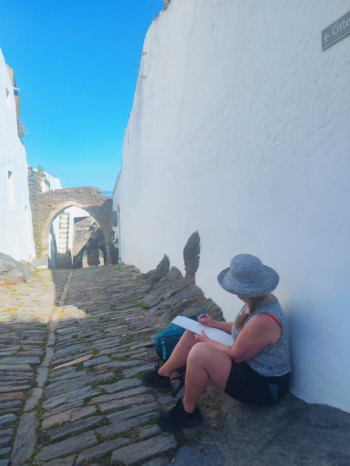 Tourist sketching in a narrow cobblestone street in Monsaraz, Portugal, with an archway in view.