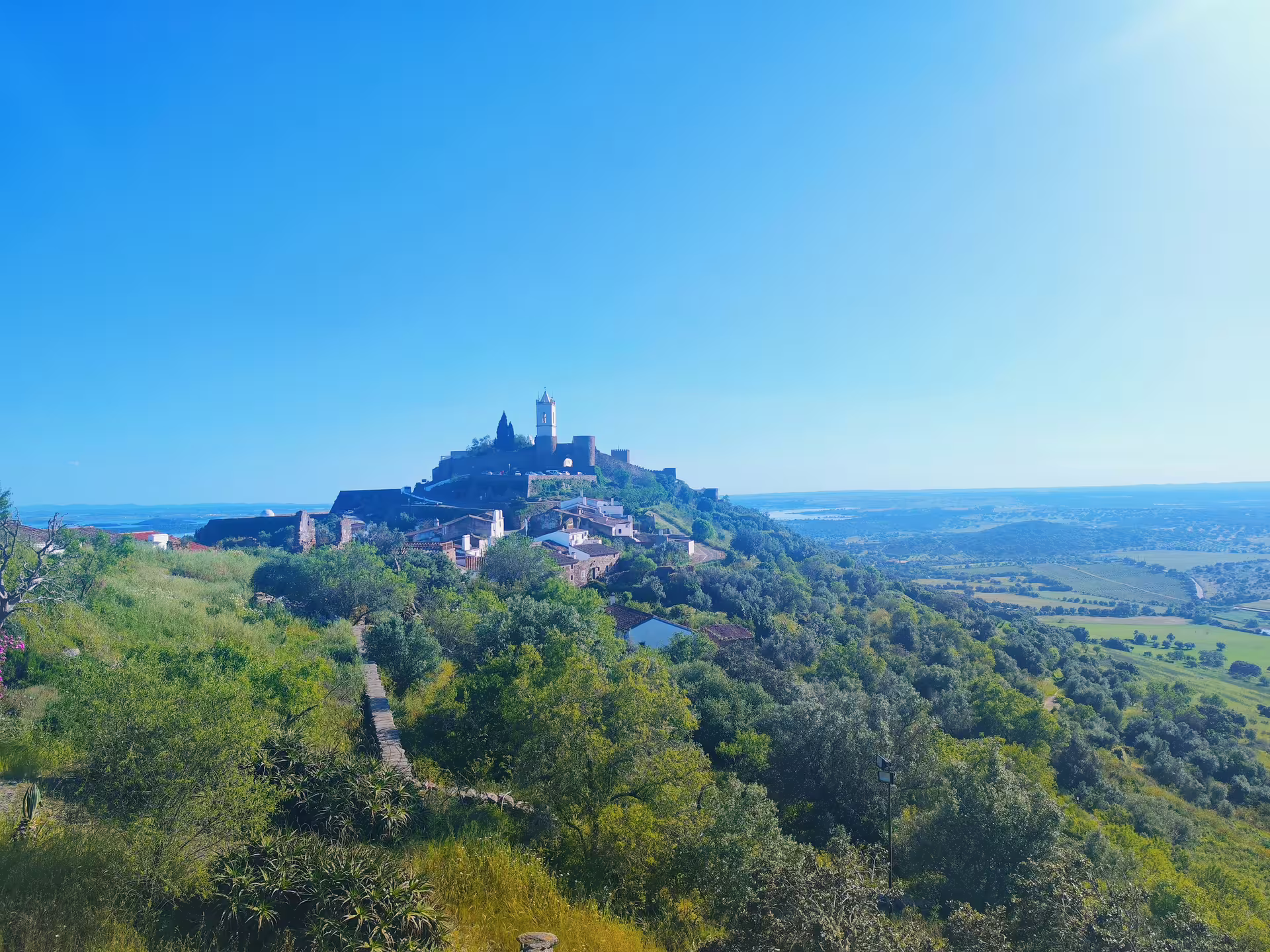 Panoramic view of Monsaraz village perched on a hilltop, surrounded by lush greenery and scenic landscape.