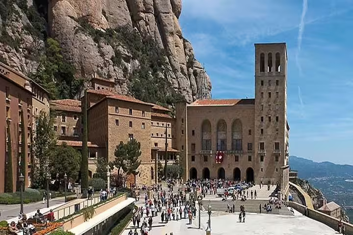Tourists gather in the vibrant courtyard of Montserrat Abbey, set against the rugged Montserrat mountain backdrop.