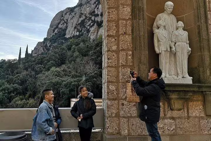 Tourists enjoy a Montserrat private tour, capturing memories near a statue with stunning mountainous backdrop and La Moreneta access.