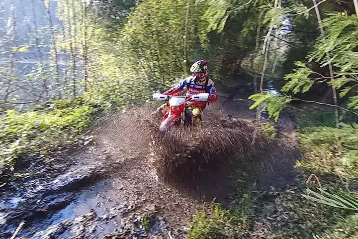 Motorcyclist splashing through a muddy trail surrounded by lush greenery on an enduro tour in Marco de Canaveses.