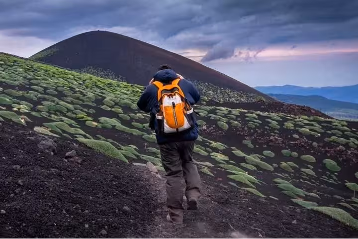 Hiker with orange backpack trekking up lush green slopes of Mount Etna on a half-day guided tour.