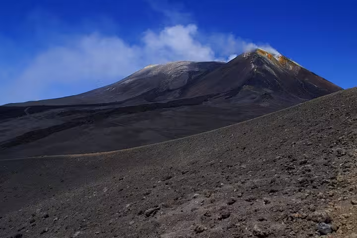 Barren, rocky terrain of Mount Etna with smoke rising from the summit against a vivid blue sky.