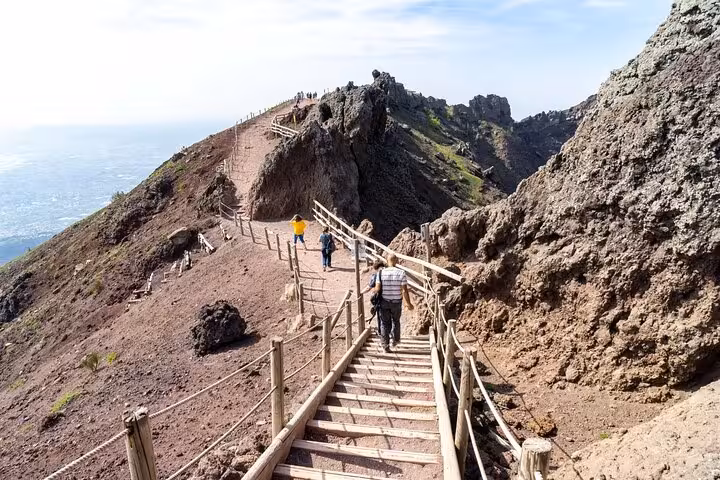 Visitors hike the scenic trail along the rugged slopes of Mount Vesuvius under a bright blue sky.