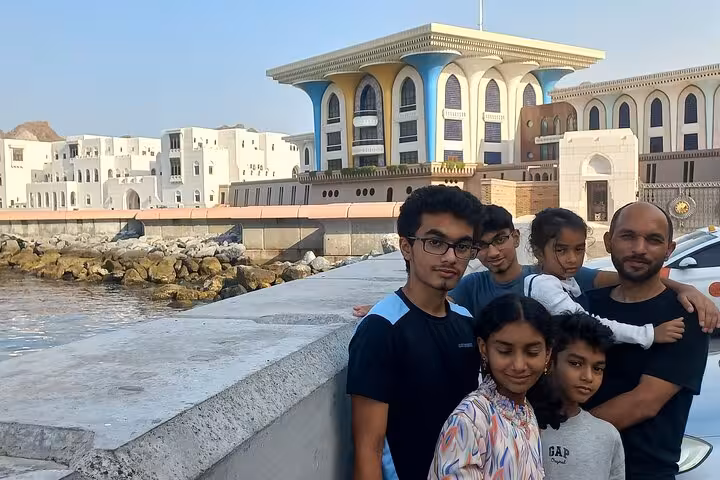 Family poses on Muttrah Corniche with Al Alam Palace in background during Muscat evening sightseeing taxi tour along waterfront