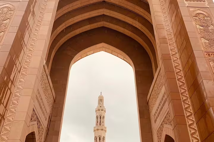 Grand Mosque's intricate arch framing a towering minaret under a cloudy sky during a private morning tour in Muscat.