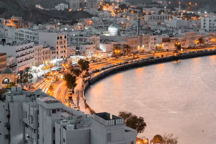 Evening view of Muttrah Corniche in Muscat with illuminated seafront road, white buildings and domed mosque on private taxi tour