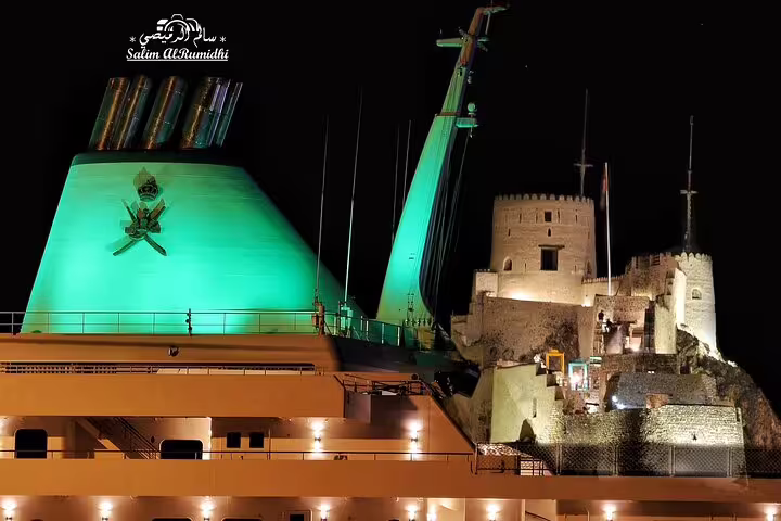 Night view of a brightly lit ship and historic fort in Muscat, highlighting the blend of modern and ancient architecture.