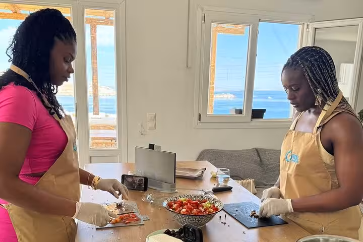 Guests chopping fresh ingredients in an authentic Mykonos cooking class with Aegean Sea views through windows