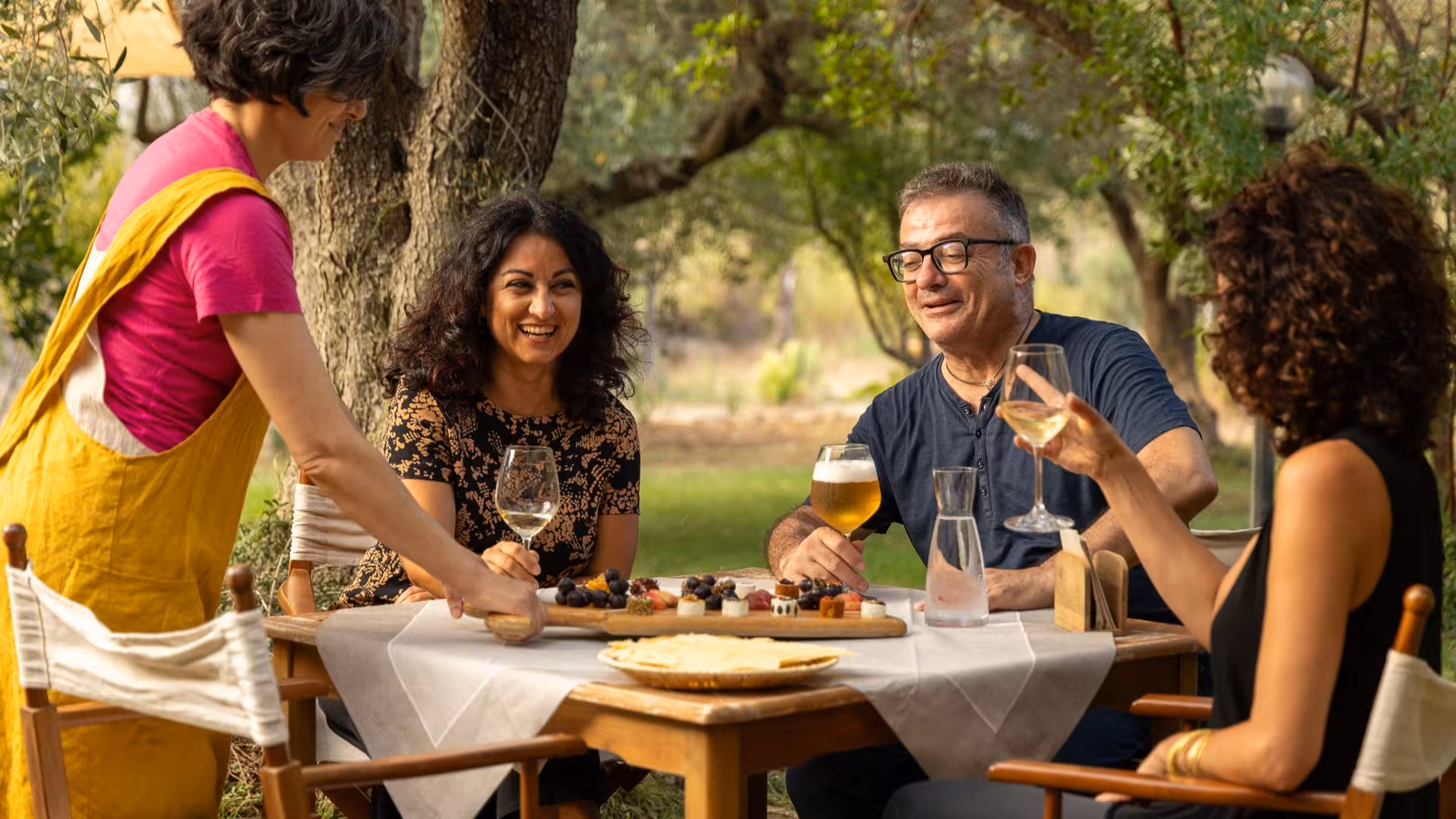 Group enjoying Oliena goat cheese tasting outdoors with wine and laughter in a scenic garden setting.