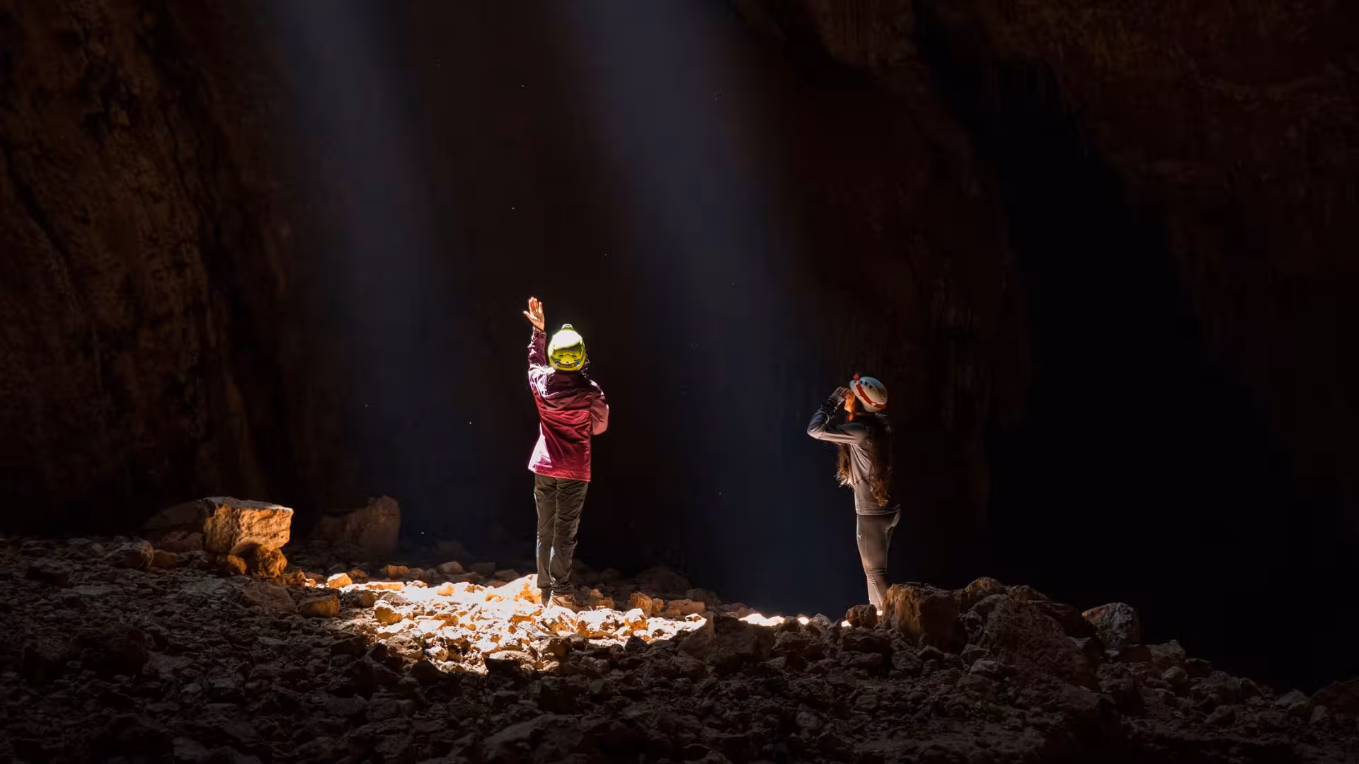 Two adventurers in helmets marvel at the sunlit interior of Sa Nurre de Su Hoda cave, Oliena.