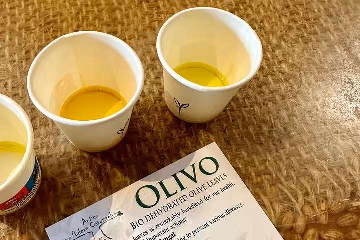 Two cups of olive oil with a brochure on benefits of olive leaves, highlighting tasting experience in Tuscany.