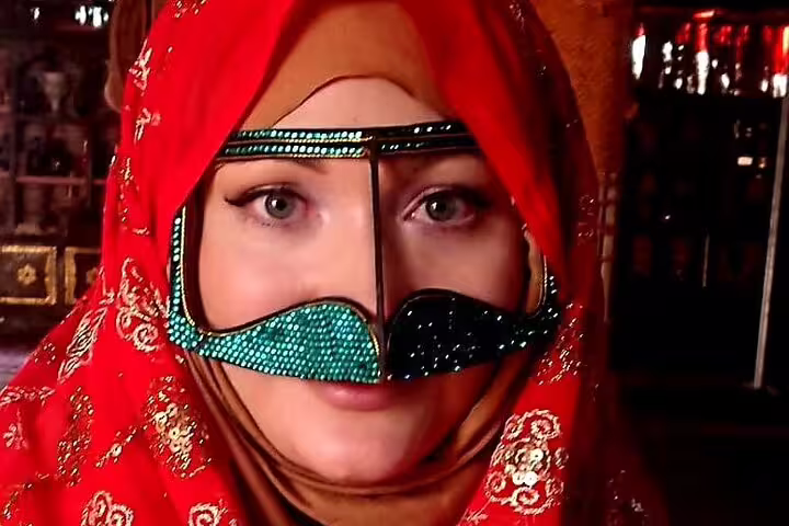 Woman in traditional Omani attire wearing a vibrant red headscarf and bejeweled face mask during desert tour.