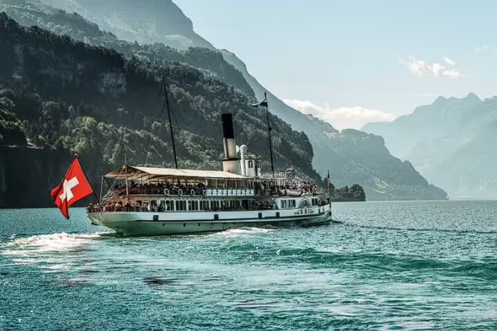 Historic paddle steamer with Swiss flag cruising through the serene waters of Lake Lucerne, bordered by mountains.