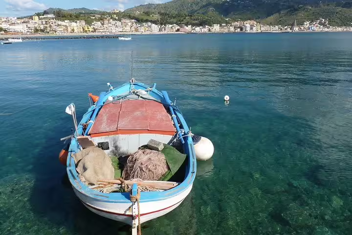 Traditional fishing boat floating on clear blue water near Giardini Naxos, with Sicilian coastal town and hills in the background