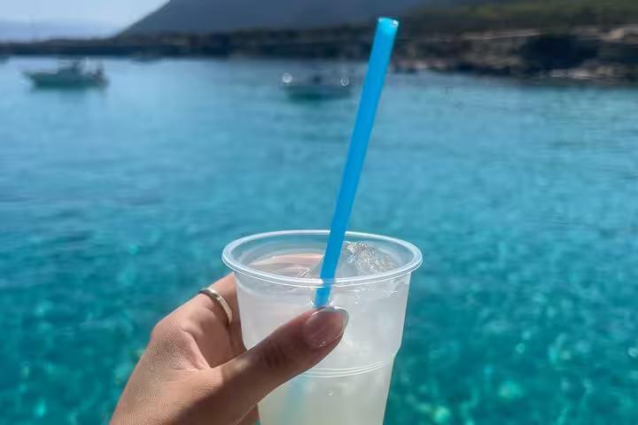 Hand holding cold drink on boat with Akamas Blue Lagoon turquoise sea in background on Paphos day trip