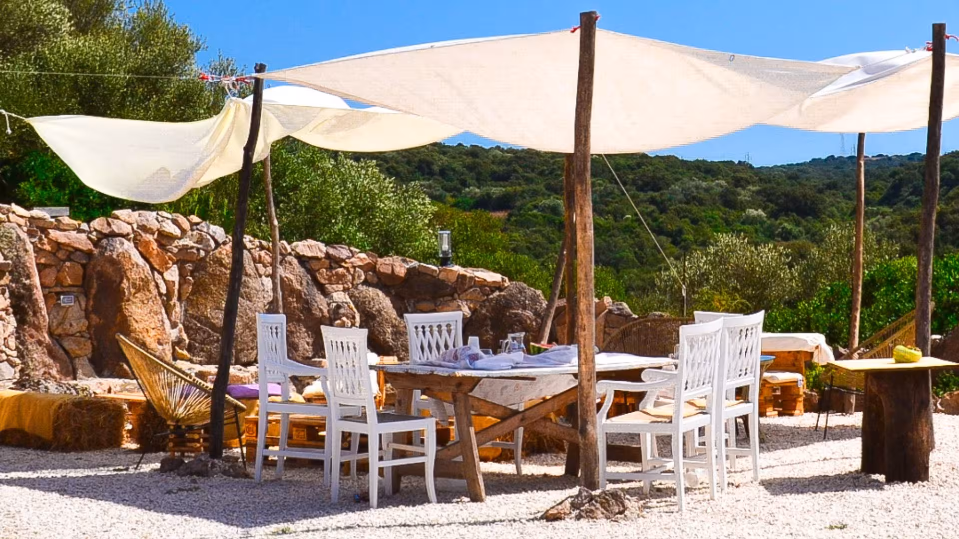 Rustic outdoor dining setup at Perfugas glamping site, featuring white chairs, wooden table, and sunshade.