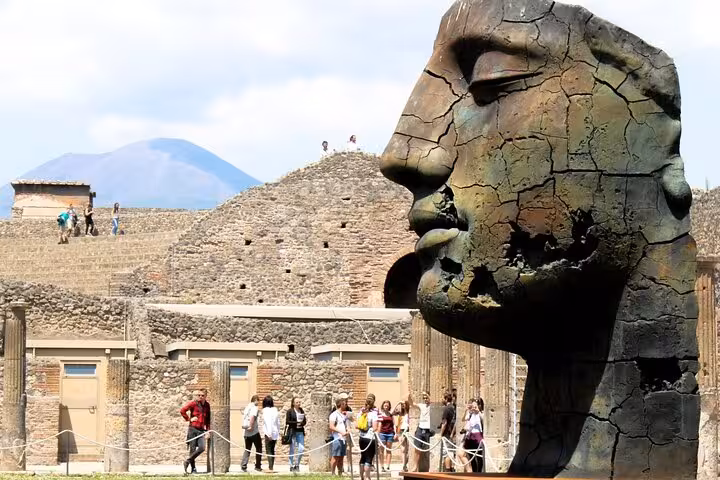 Discover Pompeii's striking bronze head sculpture against a backdrop of historic ruins and Mount Vesuvius on a guided tour.