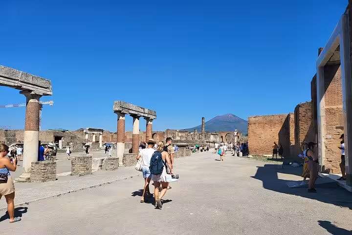 Tourists explore ancient Pompeii ruins with Mount Vesuvius in the background on a sunny day, part of guided tour from Sorrento.