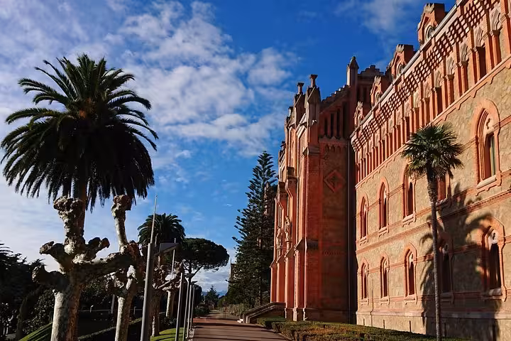 Majestic architecture of the Pontifical University in Comillas, surrounded by lush greenery and palm trees under a blue sky.