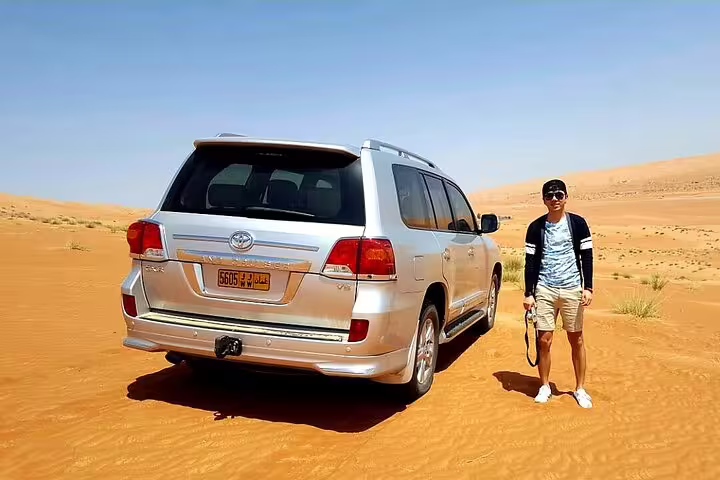 Tourist standing beside a silver SUV in Wahiba Sands, highlighting private desert transfer from Muscat experience.