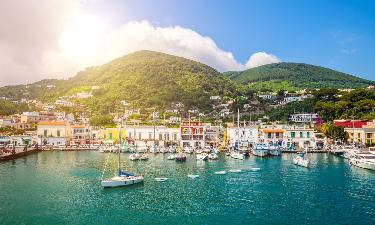 Colorful marina on Procida island with yachts moored in turquoise water beneath lush green hills on a sunny VIP catamaran day