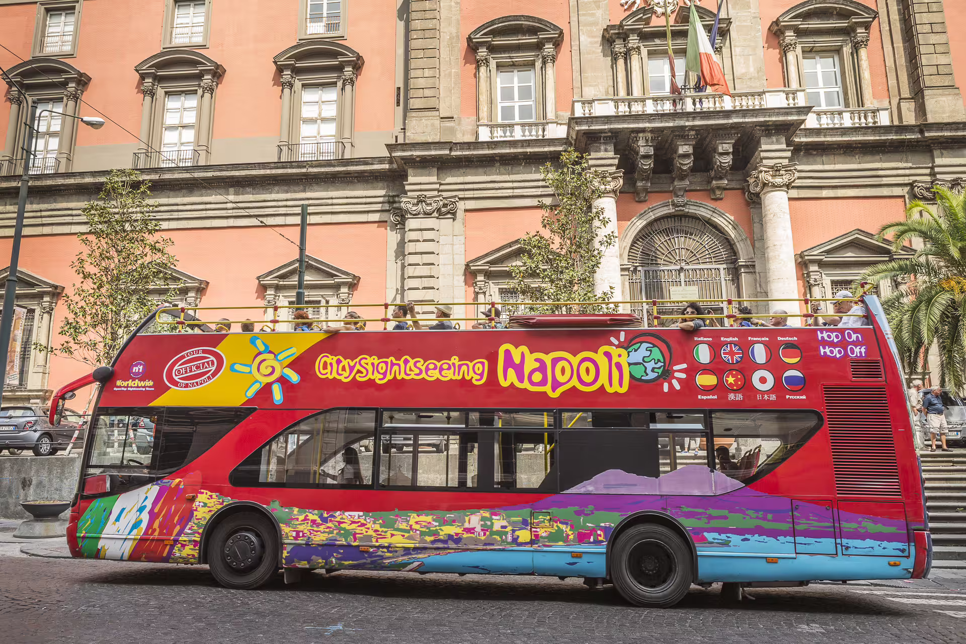 Red City Sightseeing Napoli hop on hop off bus in front of historic palace, offering open-top city tour of Naples landmarks