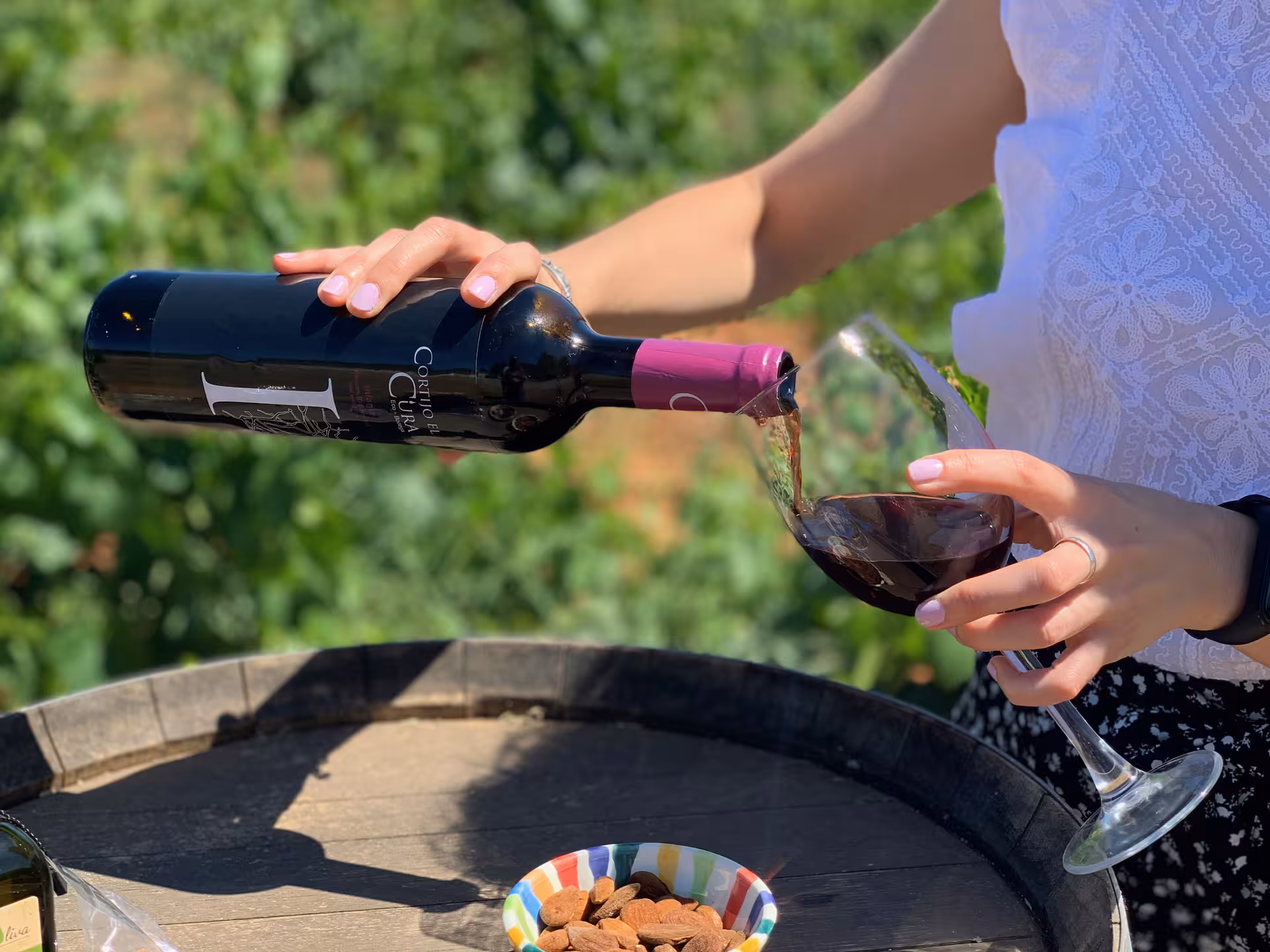 Pouring red wine into a glass during a tasting tour in Almería, with a vineyard backdrop and almonds on a barrel.