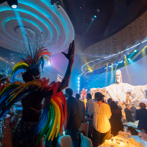 Vibrant performer with rainbow feathers during Rio By Night show at Roxy Copacabana, featuring iconic Christ statue backdrop.