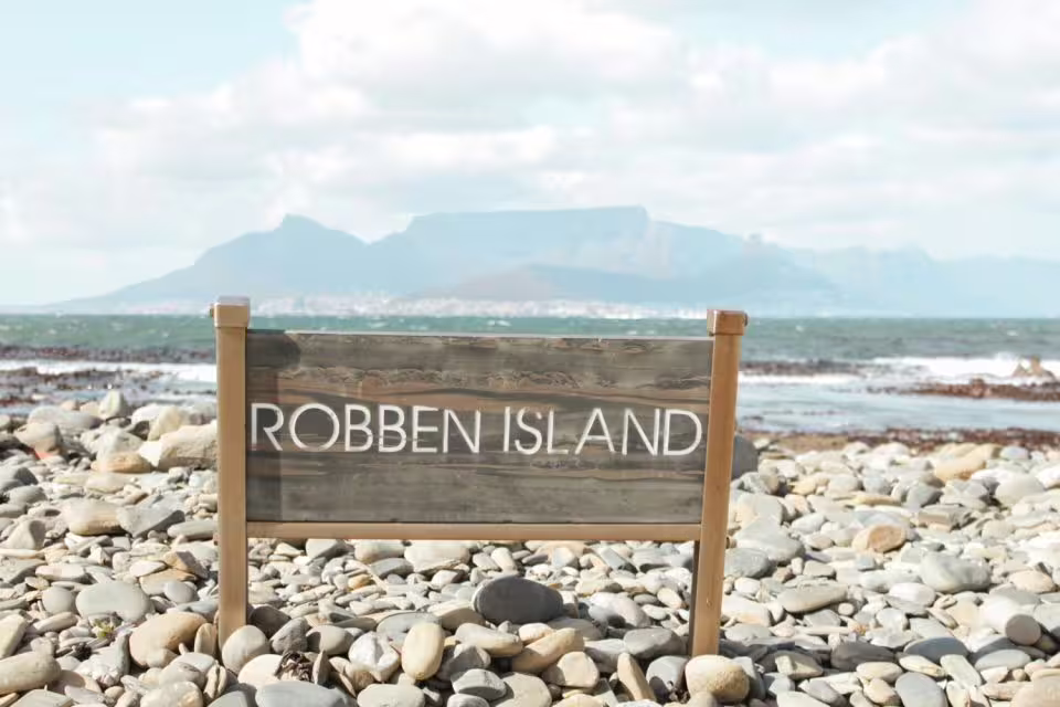 Sign for Robben Island on a rocky beach with ocean and Table Mountain in the background, highlighting a historic UNESCO site.