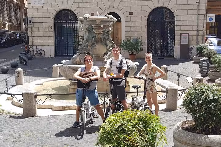 Cyclists pause by a charming Roman fountain during a small group e-bike tour, basking in sunny weather.