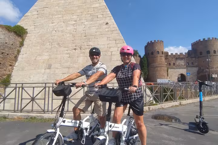 Tourists on e-bikes explore Rome's Pyramid of Cestius and ancient city walls under a clear blue sky.