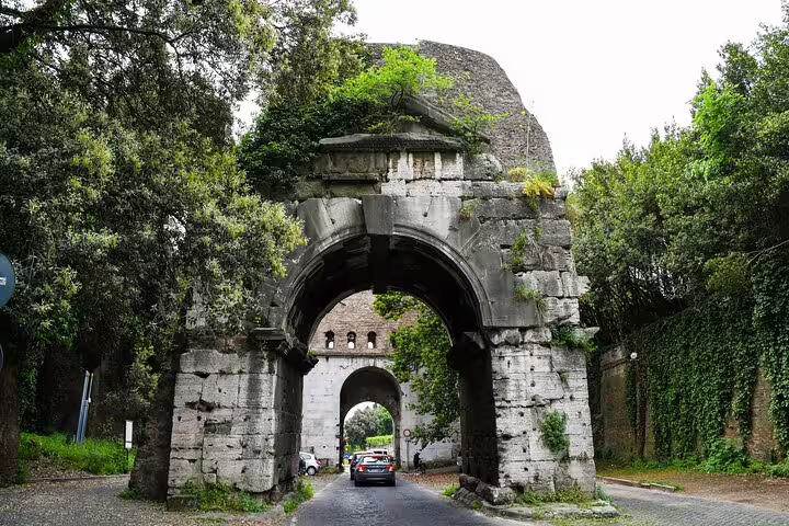 Family-friendly Rome tour passing under ancient Aurelian Walls gate with cars driving through lush green historic archway