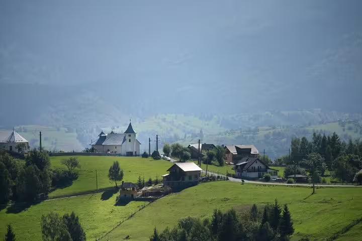 Green hillside Romanian mountain village with white church and traditional houses in misty Carpathian landscape